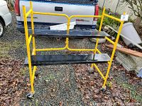 Metal scaffold frame painted yellow with two black removable platforms on it, outdoors on gravel surface.