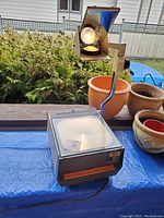 Side view of overhead projector on blue tarp with surrounding planters and outdoor setting. Shows lamp lit inside projector head and frosted glass projection surface.