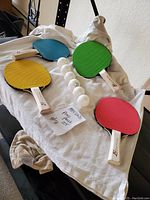 Photo showing four colorful East Point ping pong paddles in blue, green, yellow, and red with several white ping pong balls arranged on a white cloth.