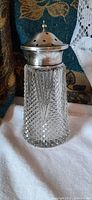Sugar caster with cut crystal glass body and a silver cap with holes for sprinkling, sitting on fabric background.