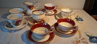 Six teacups with matching saucers arranged with a creamer and sugar bowl on a white tablecloth with embroidered details. Various floral and geometric designs with gold trims visible.