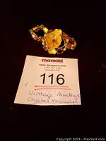 View of the gold tone floral bracelet with large central flower and smaller flowers accented by amethyst-colored crystals, laid beside auction lot number card.