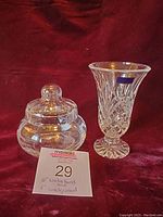 Two glass items displayed on a red fabric: a clear crystal vase with label and a round lidded glass jar.