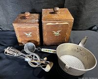 Two wooden canisters with golden eagle on front, vintage hand mixer, strainer, and ladle laid out on black background.