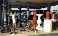 Photo showing a variety of candlestick holders made from wood, ceramic, brass, copper, and metal, along with several candles including tapers and pillar candles, placed together on shelving.
