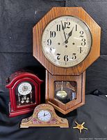 Overview photo of three clocks: large oak wall clock, floral quartz mantel clock, small red mantel clock, all on black cloth background.