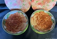 Two porcelain dessert plates with large peach and pink rose floral designs and a decorative green rim, shown with floral round display boxes labeled 'Sitting Pretty'.