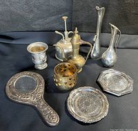 Photo of full lot items on black fabric: a silver plated hand mirror, small goblet, brass oil can/plant mister, two pewter bud vases, gold-tone votive holder, silver plated octagonal tray, and small round dish.