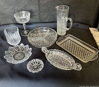 Overview of the eight vintage clear glass items: daisy trinket dish, petal plate, lowball whiskey glass, dessert dish, three-section condiment dish, pickle dish, relish tray, and pint pitcher on dark backdrop.