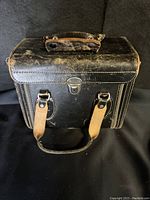 Front view of black vintage leather camera case showing handle, metal clasp, and two tan leather straps.