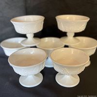 Seven milk glass pedestal compote bowls shown front, arranged with three elevated in background and four in foreground on flat surface to highlight grapevine detail.