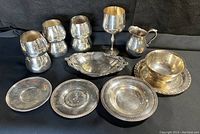 Full display of vintage silver plated collection showing cups, goblet, trays, pitcher, and bowl on black background.