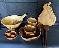 Full view of wooden bowls, nested plates, small bowls, serving spoons, salt and pepper shakers, and collapsible pear-shaped trivet/bowl on black background.