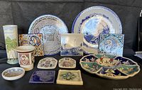 Full assortment of 15 assorted plates, plaques, tiles, and ceramic decorative items arranged closely on a black surface, showing differentiated styles and colors including blue and white motifs and colorful floral patterns.