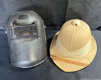 Welding mask and pith helmet side by side on black background.