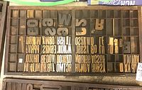 Wooden flat divided into compartments holding various wooden letter and number type blocks in assorted sizes and fonts.
