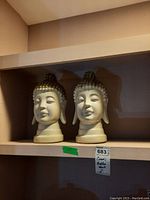 Photo showing two ceramic Buddha head sculptures on a shelf with label and measurement.