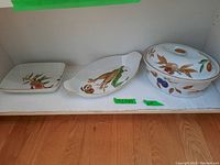 Photo showing all three ceramic serving pieces together on a shelf: oval covered serving dish and two serving plates with Evesham fruit and leaf designs and gold trim.