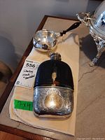 Flask and sauce pan displayed together on beige cloth atop table.
