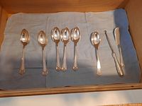 Six spoons, butter knife/cutter, and sugar hook arranged on cloth in drawer