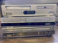 Three silver and black VCR/DVD combo units stacked, showing front side with DVD and VCR slots, buttons and ports.