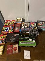 Photo of various card games grouped together on a floor including Giant UNO, standard UNO, Exploding Kittens, Fluxx, Sheeetz Beanz, Jumbo Playing Cards, The Oregon Trail, Phase 10, and Taco vs Burrito