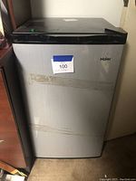Closed front view of the Haier mini fridge showing tape and sticker residue on the silver door with black top and sides.