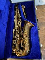 Brass tenor saxophone inside a blue velvet-lined case with visible tarnish and wear.