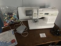 Full view of Brother SQ9185 sewing machine with foot pedal, power cord, instruction manual, and bag of colorful thread spools on wooden table.