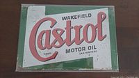 Photo of a rectangular metal Castrol Wakefield Motor Oil advertising sign with red lettering on white and green background, some slight wear visible.