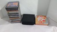 Stack of 21 jewel case CDs, black soft CD holder for 23 loose CDs, and 'Hits of the 70s' tin box