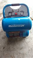 Front view of blue Mastercraft air compressor showing brand logo on tank and control panel with gauges and knob.