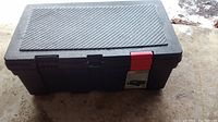 Closed black rectangular plastic storage box with textured lid and one red latch on the front; other front latch missing.