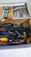 Top view of box containing saw blades in packaging, measuring tape, and various hand tools including pliers and screwdrivers with plastic and rubber handles in assorted colors