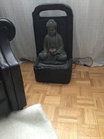 Front view showing the statue in seated Buddha meditation pose, holding a lotus flower with power cord visible on floor near base.