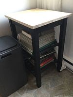 Front angle showing butcher block top, black frame base with two shelves containing books and magazines.