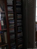 Photo showing two tall wooden CD towers filled with vintage CDs, placed next to bookshelf and window with partial natural light.