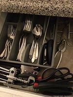 Stainless steel cutlery including forks, spoons, butter knives, kitchen scissors, corkscrew, and garlic press organized in a plastic tray