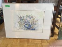 Front view of framed watercolour print showing floral arrangement in blue vase, with white matting and silver frame, resting on parquet floor.