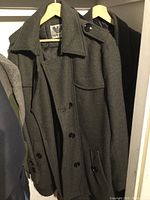 Dark gray wool overcoat hanging on a wooden hanger inside a closet, buttons visible and classic collar spread open.