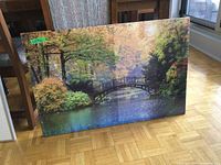 Photo of fall themed river and bridge canvas art leaning against dining chairs on wood parquet floor. Shows entire canvas with details of colors and reflections.
