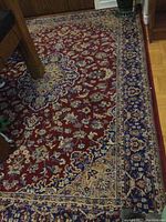 Close-up photo of the red Persian style area rug showing floral pattern near a wooden table leg on hardwood floor.