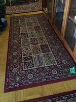 Photo showing one Persian style runner rug on parquet floor near glass cabinet. Rug features patchwork rectangular panels of floral and geometric patterns with red floral border.