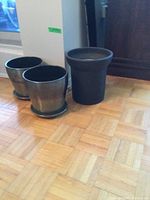 Three plant pots on a wooden floor near a wall and door, two metallic-colored with saucers and one black matte larger pot