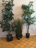 Front view of five artificial plants showing one broad-leaf tall tree in a square base on the left, three medium grassy and leafy plants in round pots in the middle, and one variegated leaf tall tree in a round base on the right, all standing on parquet flooring against white wall.