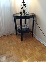 Two black round side tables with glass tops and lower shelves placed next to walls in a room with wooden parquet flooring.