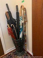 Photo showing vintage metal umbrella holder filled with various umbrellas and a decorated wooden walking stick.