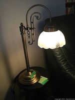 Vintage brass desk lamp with white frosted scalloped glass shade illuminated, showing detailed brass scrollwork and patterned glass.