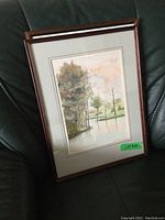 Framed watercolour landscape print showing trees and water, propped on green leather couch.