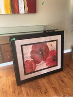 The framed print leaning against a wall showing the full 27x21 inch frame with three red poppies in the artwork.